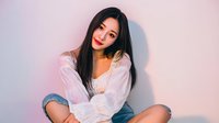 Young Asian woman with long dark hair wearing white blouse and denim shorts poses against pastel background with soft pink lighting.