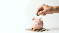 Hand dropping coin into pink piggy bank surrounded by scattered coins on white background for savings and financial planning concepts.