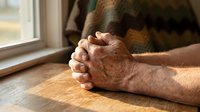 Senior man with weathered hands clasped together on wooden table near window, expressing contemplation, prayer, or emotional moment. Suitable for healthcare, spirituality, aging.