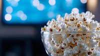 Fresh popcorn overflowing in glass bowl against blurred blue bokeh background, creating cozy movie night atmosphere for entertainment and snacking.