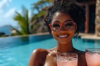 Young African American woman with natural curly hair enjoying tropical vacation by infinity pool, wearing round sunglasses and holding pink cocktail, radiating joy.