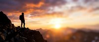 Silhouette of hiker standing on rocky mountain peak at golden sunset, watching expansive landscape view. Adventure and exploration concept for travel and outdoor lifestyle projects.