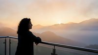 Woman with long dark curly hair wearing black jacket gazing at misty mountain landscape at sunrise from modern balcony railing.