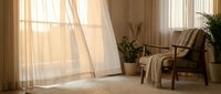 Cozy living room corner with wicker armchair, soft throw blanket, potted plants and flowing curtains in warm natural sunlight for home decor inspiration.