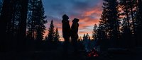 Young couple standing together by campfire in forest at sunset, silhouetted against dramatic orange and pink sky surrounded by tall pine trees, romantic outdoor camping scene.