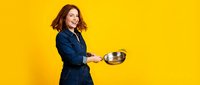 Young Caucasian woman in blue denim jacket holding metal bowl on bright yellow background, smiling and looking at camera in studio portrait.