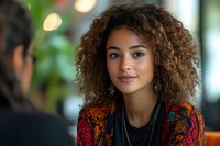 Young mixed race woman with natural curly hair wearing colorful patterned sweater smiling during casual conversation at cafe, soft bokeh background.