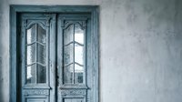 Vintage weathered blue wooden doors with ornate glass panels against textured gray wall creating rustic Mediterranean architectural detail for interior design.