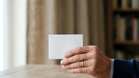Mature man with light skin tone holding blank white card in hand, wearing dark blue sleeve with white checkered cuff, indoor setting with curtains and bookshelf background for business communication.