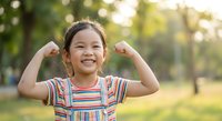 Happy Asian girl child flexing muscles showing strength and confidence outdoors in sunny park with natural bokeh background for health and wellness concepts.