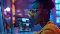 Young Black woman with braids and glasses working at computer in vibrant neon-lit tech workspace, focused and professional.
