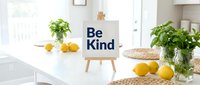 Wooden easel with Be Kind sign on white kitchen counter, surrounded by fresh lemons and green plants in bright, airy home interior.