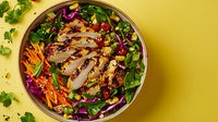 Fresh grilled chicken breast sliced over vibrant mixed salad with purple cabbage, carrots, chickpeas and cilantro in round bowl on yellow background.