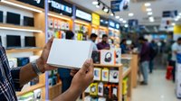 Male customer of South Asian descent holding blank white box in electronics retail store with product displays and shoppers in background.