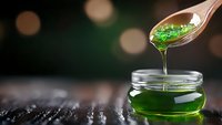 Green transparent syrup dripping from wooden spoon into glass jar on dark wooden surface against blurred bokeh background, macro food photography.