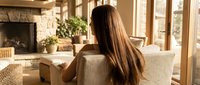 Young woman with long brown hair relaxing on beige sofa in modern living room with fireplace and large windows. Cozy home interior lifestyle concept.