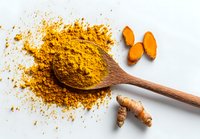 Ground turmeric powder on wooden spoon with fresh turmeric root slices on white background, popular spice for cooking and natural health remedies.