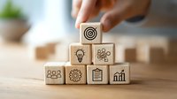 Hand arranging wooden blocks with business icons showing target, lightbulb, gears, people, clipboard, and growth chart on wooden surface.