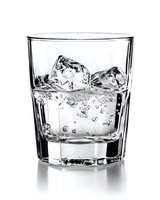 Glass of sparkling water with ice cubes on white background, bubbles rising through clear liquid in classic tumbler, high contrast black and white photo.