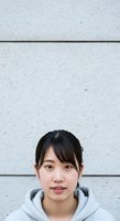 Young East Asian woman with long dark hair and bangs wearing gray hoodie against light concrete wall with horizontal seams, suitable for portrait and lifestyle content.