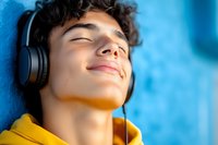 Young Hispanic man in yellow hoodie listening to music with wireless headphones, eyes closed with peaceful expression against blue background.