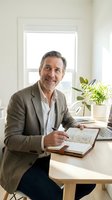 Middle-aged man with gray hair and beard, Caucasian ethnicity, wearing olive blazer and light blue shirt, sitting at wooden desk writing in notebook with pen. Bright modern office with potted plant, laptop, and large window with natural light. Professional businessman or entrepreneur working on planning and organization tasks.