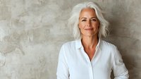 Confident mature woman with silver hair wearing white button-up shirt standing against textured wall background, representing success and professional lifestyle.