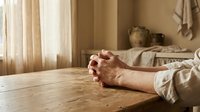 Elderly person hands clasped in prayer on wooden table in warm sunlit room with peaceful atmosphere for meditation and spiritual reflection.