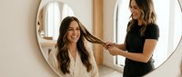 Young brunette woman getting hair styled by professional hairstylist in modern salon with round mirror and natural lighting for beauty services.