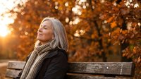 Mature blonde woman relaxing on park bench during golden autumn sunset, enjoying peaceful moment outdoors with warm sunlight and colorful fall foliage.