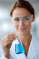 Young female scientist in protective eyewear and lab coat holding blue liquid in flask, smiling confidently while conducting research in laboratory setting.