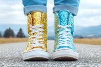 Mismatched glitter high-top sneakers in gold and turquoise with white laces and soles, worn with rolled blue jeans on asphalt road against blurred nature background.