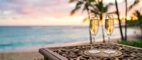 Two champagne glasses on tropical beach table at sunset with palm trees and ocean view for romantic celebration and luxury vacation moments.