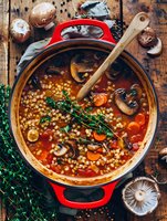Homemade mushroom and pearl couscous soup with fresh thyme, carrots, and tomato broth in red cast iron pot on rustic wooden background, overhead view.