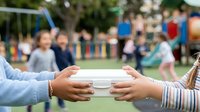 Young child with medium skin tone handing over a white plate or container to another child at an outdoor community event or school gathering, with blurred playground and participants in background.