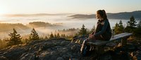 Woman with dark hair and medium complexion sitting on rocky outcrop overlooking misty forested valley at sunrise, wearing dark jacket and backpack, contemplating scenic mountain landscape with evergreen trees and fog.