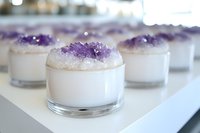 Purple amethyst crystal clusters adorning white soy candles in glass holders create serene atmosphere for meditation, spa treatments and spiritual practices.