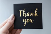 Gold calligraphy thank you text on black card held by feminine hand with neutral manicure against light background, elegant minimalist gratitude message for special occasions.