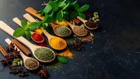 Colorful spices and herbs in wooden spoons on dark slate background. Fresh mint, turmeric, paprika, coriander seeds create aromatic culinary composition.