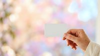 Hand holding blank white card against soft pastel blurred floral background with pink and blue tones, suitable for greeting cards, invitations, and promotional messaging.
