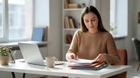 Young Caucasian woman reading documents at home office desk with laptop and coffee cup, focused on paperwork in bright modern workspace.