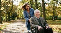 Happy senior Caucasian man in wheelchair with caring middle-aged woman caregiver in autumn park setting for healthcare and family support concepts.