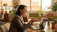 Young Asian businesswoman laughing during video call on laptop in bright modern office with plants, remote work communication and online meeting.