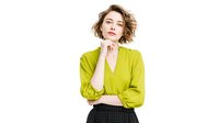 Young woman with short wavy hair wearing bright lime green blouse posing thoughtfully with hand under chin against white background.
