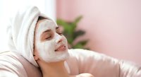 Young woman relaxing with white facial mask and towel on head in spa setting. Self-care skincare routine at home for healthy glowing skin.