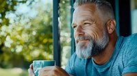 Mature bearded man with gray hair enjoying morning coffee outdoors, wearing blue t-shirt. Natural sunlight and bokeh background create peaceful atmosphere.