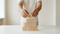 Adult male with light skin tone wearing a cream-colored shirt carefully placing a wooden block on top of a pyramid stack. Represents business growth, strategic planning, risk management, and organizational development concepts.
