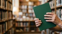 Adult male with light skin tone holding closed green hardcover book in library setting with blurred bookshelves background. Suitable for education, literacy, reading, and knowledge concepts.