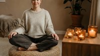 Young Caucasian woman in cozy sweater practicing meditation in lotus position on wicker cushion surrounded by warm candlelight in peaceful home interior.