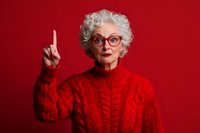 Senior woman with gray curly hair wearing red knitted sweater and stylish glasses raising index finger against vibrant red background, expressing wisdom and attention.